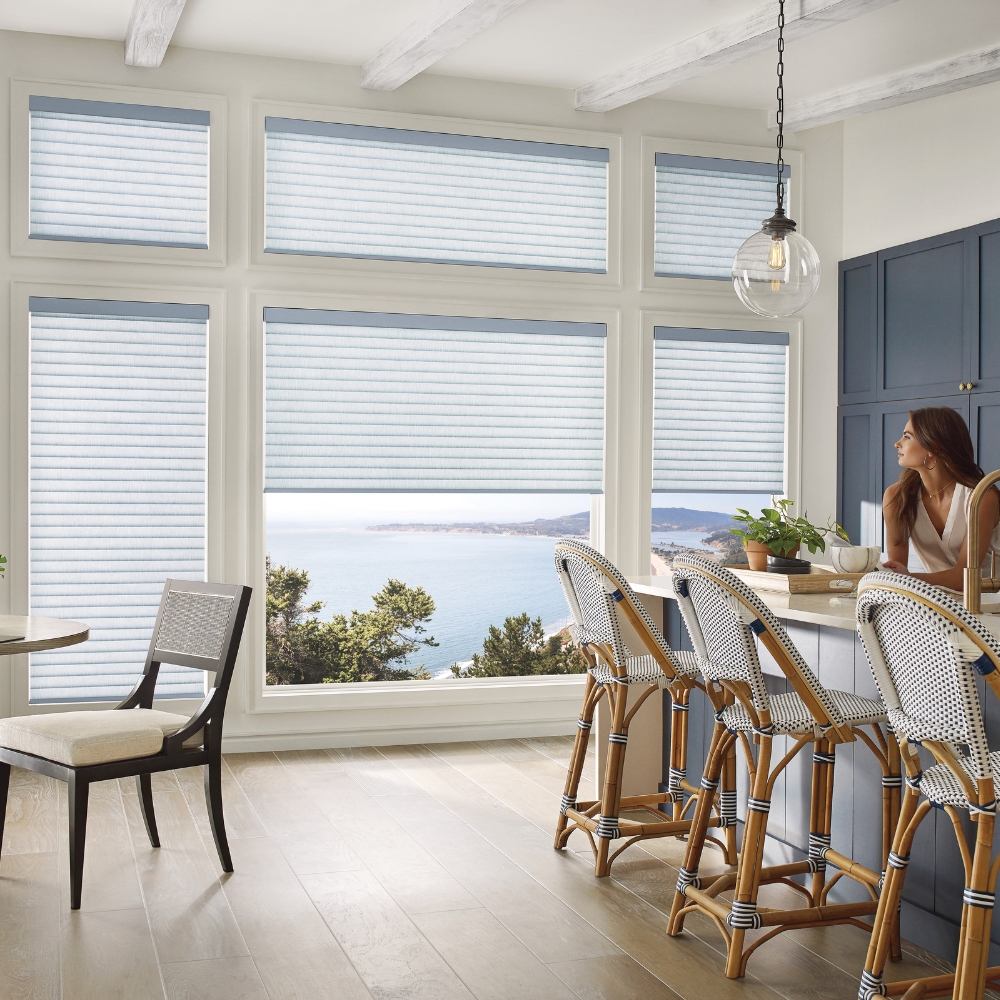 White cellular roller shades with a blue tint 2 shades are 60% open with a woman at eating bar looking at ocean view