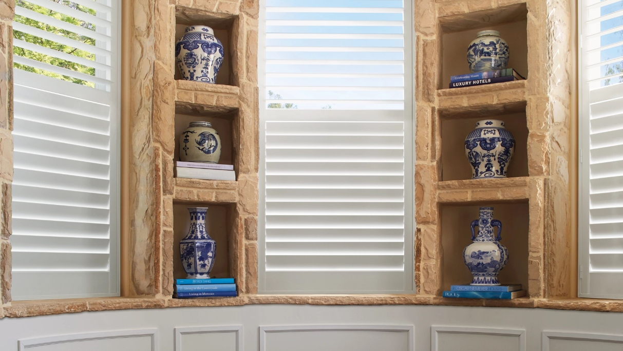 Half closed White interior shutters on 3 windows with shelves holding vases, view of trees and blue sky