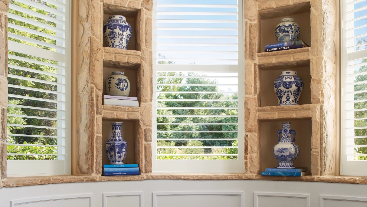 Open White interior shutters on 3 windows with shelves holding vases, view of trees and blue sky