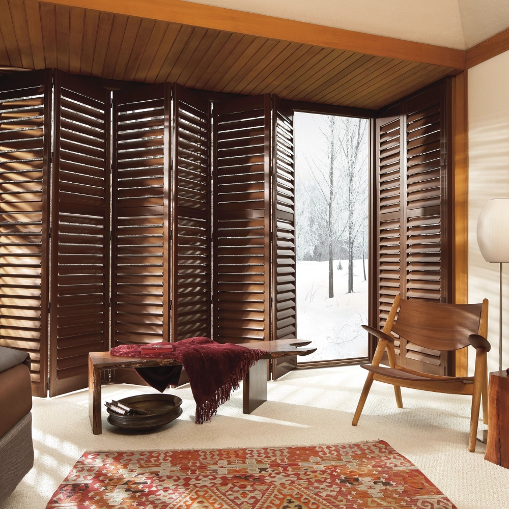 brown composite shutters bi-folding and stacking from right to left, patio door shutter 80% open with chair and table, snow and trees through patio door