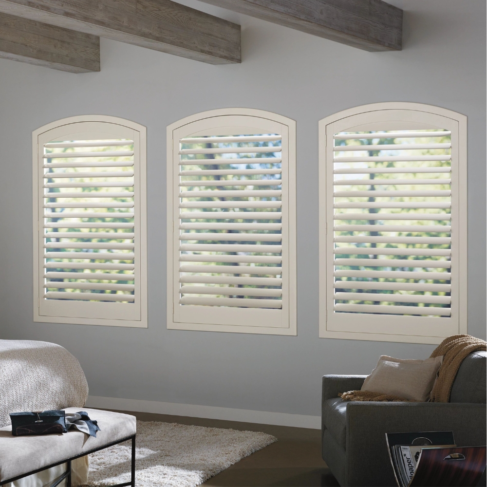 White composite shutters open on 3 windows with shallow arches in bedroom with chair, bed and bench